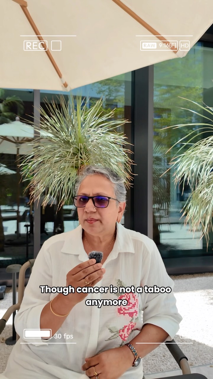 Shabnam Masood, grey-haired, in tinted glasses and a white embroidered shirt, speaking into a small microphone in a sunny courtyard. The subtitle reads “Though cancer is not a taboo anymore”.