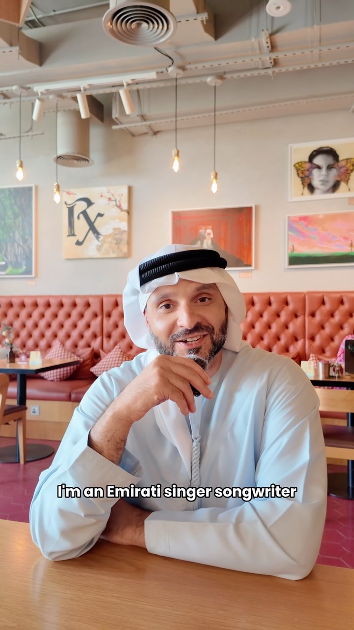 Mohamed Al Zarooni in a white kandura and ghutra, smiling at a café table with paintings on the wall behind him. The subtitle reads “I’m an Emirati singer songwriter”.